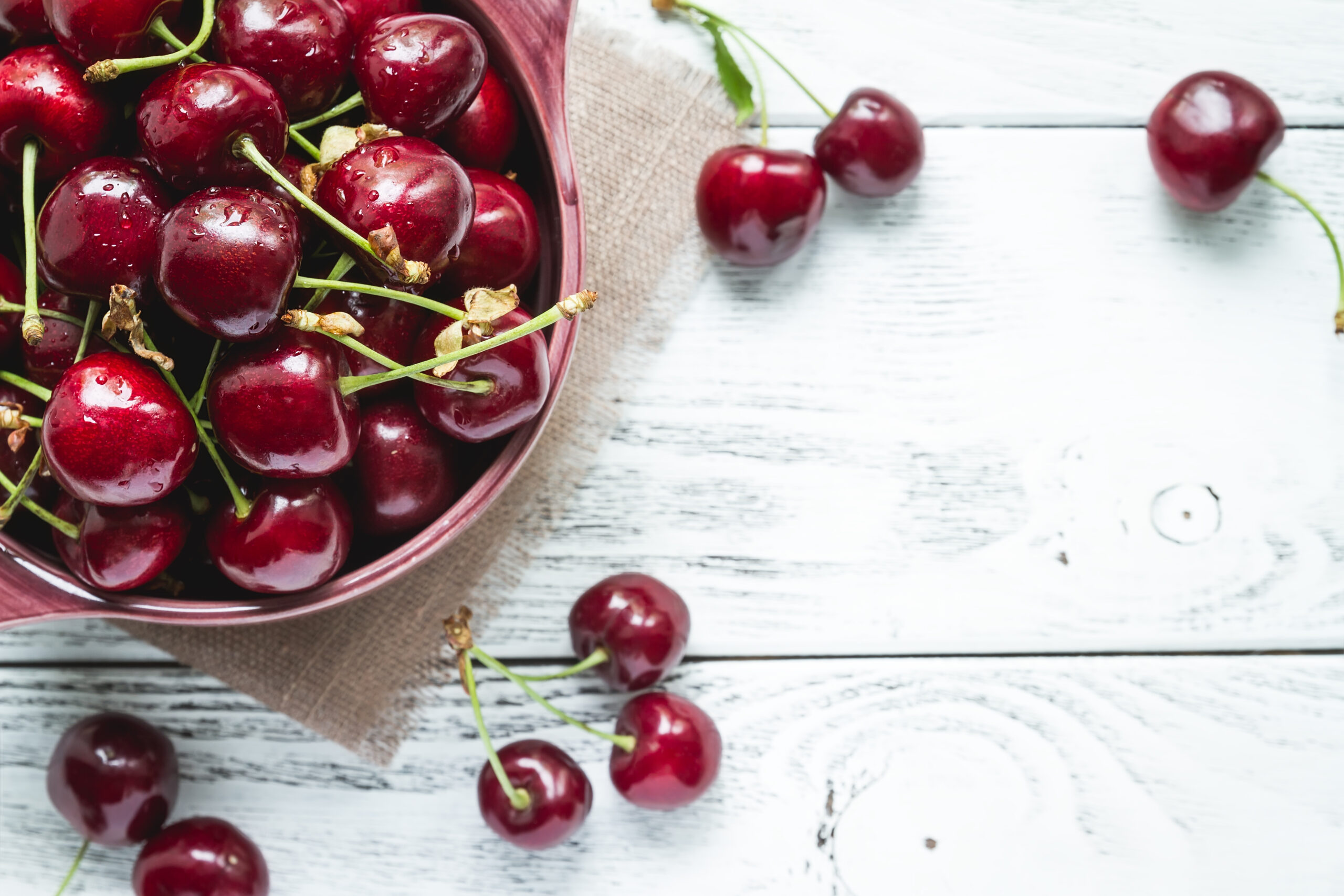 Ripe cherries in a bowl and scattered on a white wooden table.