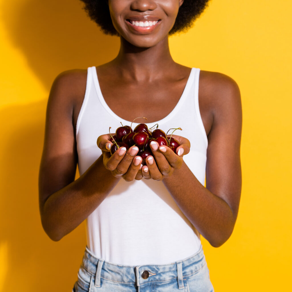 Cropped view portrait of pretty cheerful wavy haired girl holding in hands pile cherries isolated on bright yellow color background